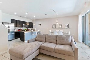 A modern living room with a beige sectional sofa, a kitchen with dark cabinets and stainless steel appliances, and a dining area with a white table and chairs.