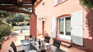 En udendørs terrasse med et spisebord, omgivet af stole, samt et overdækket siddeområde.