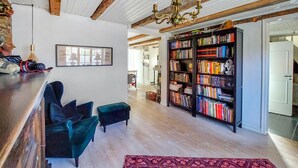 Ein geräumiges Wohnzimmer mit Holzbalkendecke, einem großen Bücherregal, zwei dunkelblauen Sesseln und einem roten Teppich mit Muster.