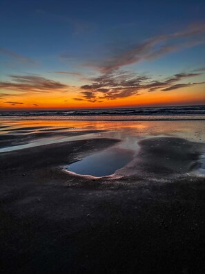 A beach with a reflective puddle and a sunset in the background.