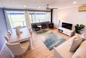 A modern living room with a dining area, a fireplace, a television, and a large window with curtains.