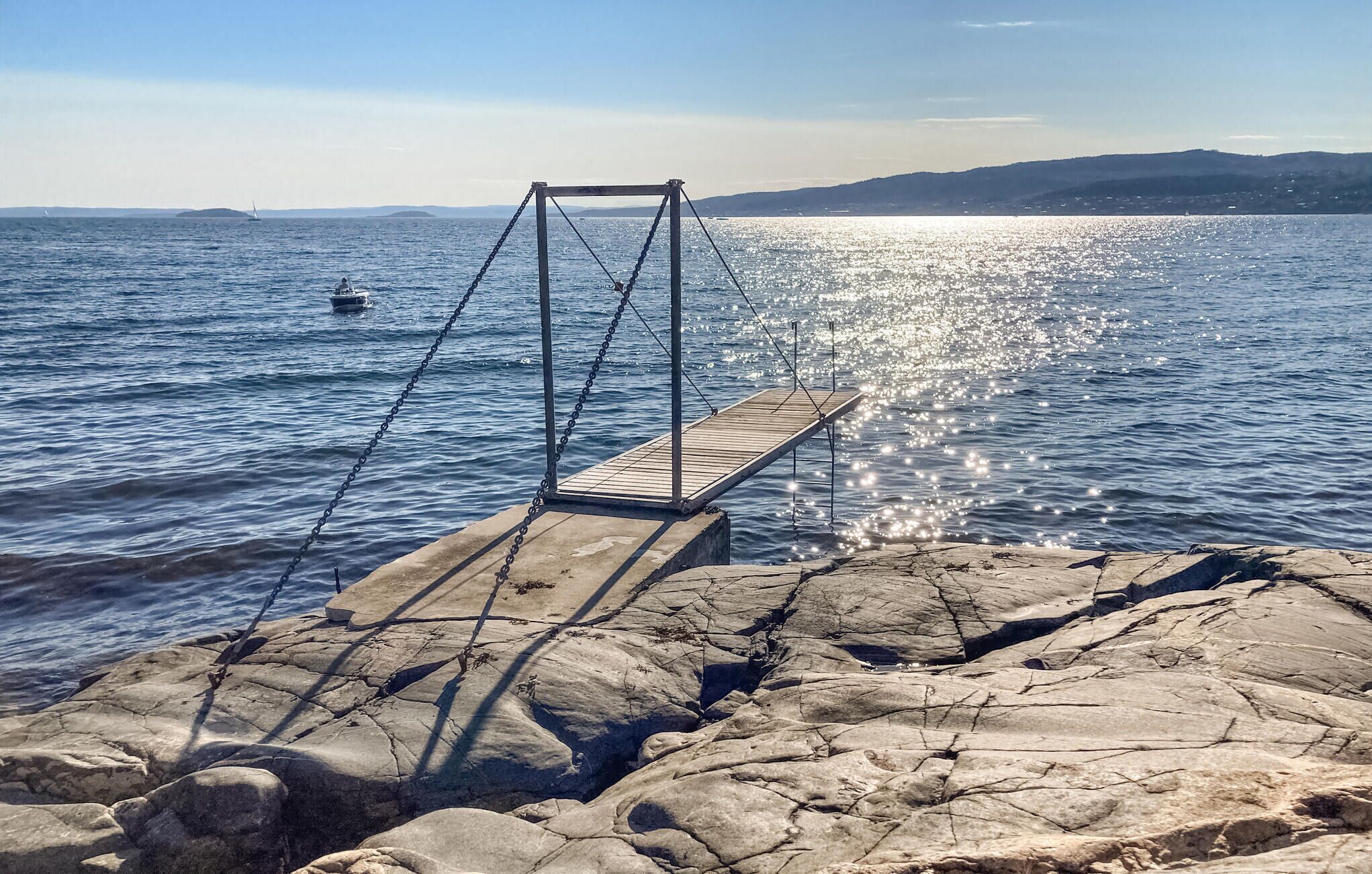 A wooden swing bridge over a rocky shoreline with a view of the sea and distant hills.