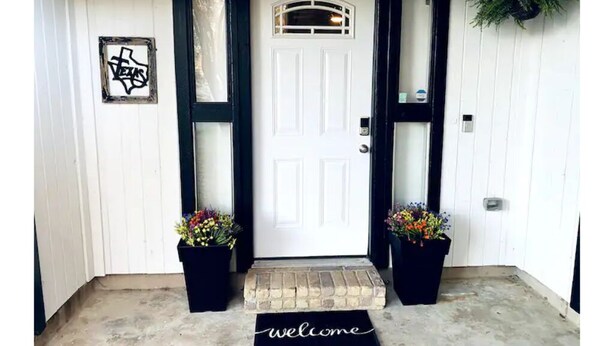 Une porte blanche avec un cadre noir, un paillasson et deux plantes en pot de chaque côté.