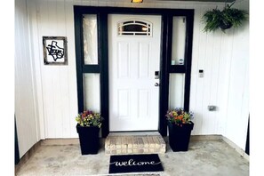 A white door with a black frame, a welcome mat, and two potted plants on either side.