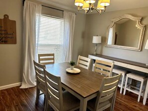 A dining area with a wooden table, white chairs, a chandelier, a mirror, and a window with blinds. - Spacious 3 BDRM for 9 - SA & NB (Live Oak)