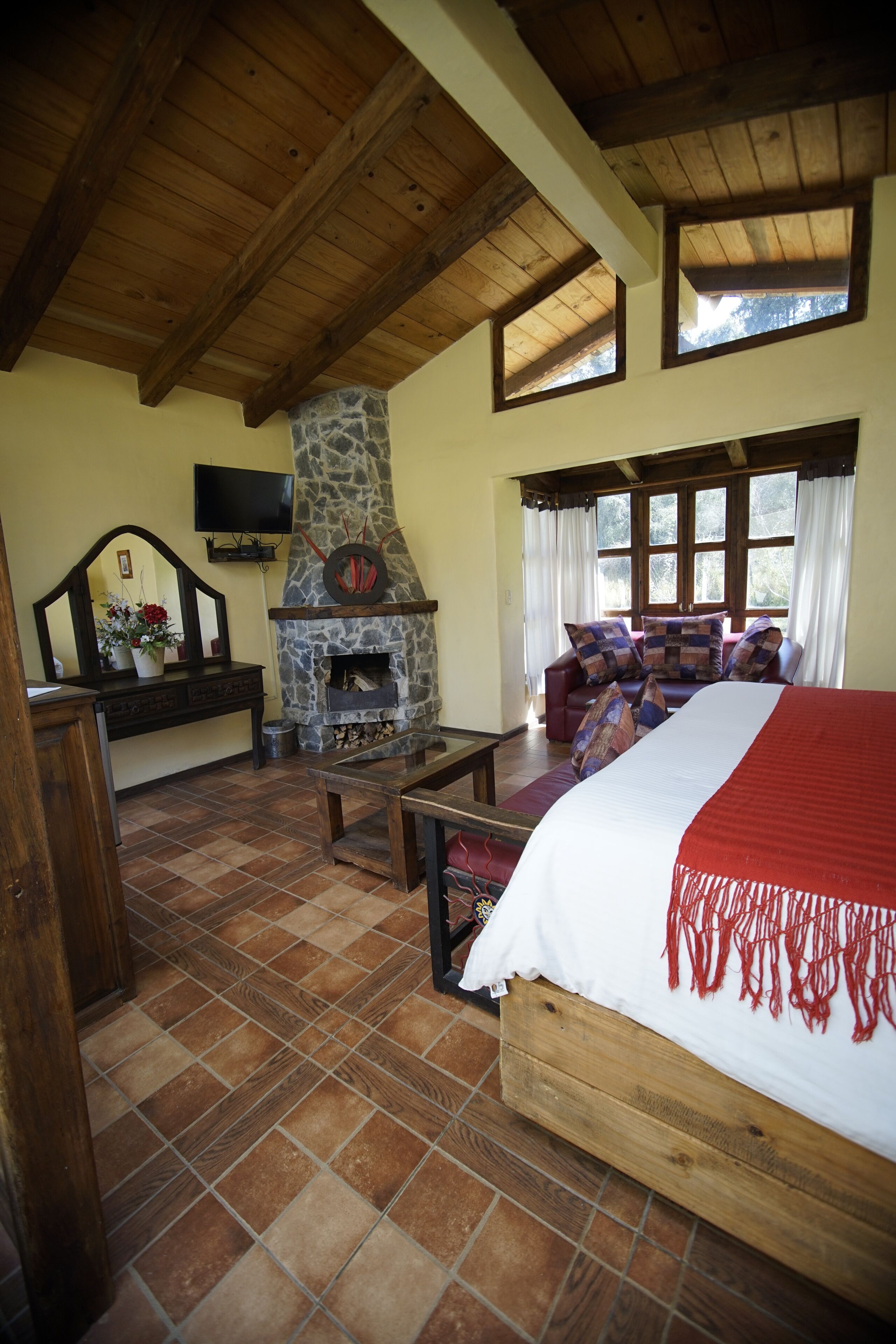 Una habitación acogedora con chimenea de piedra, una cama con manta roja, un sofá con cojines y un televisor montado en la pared.