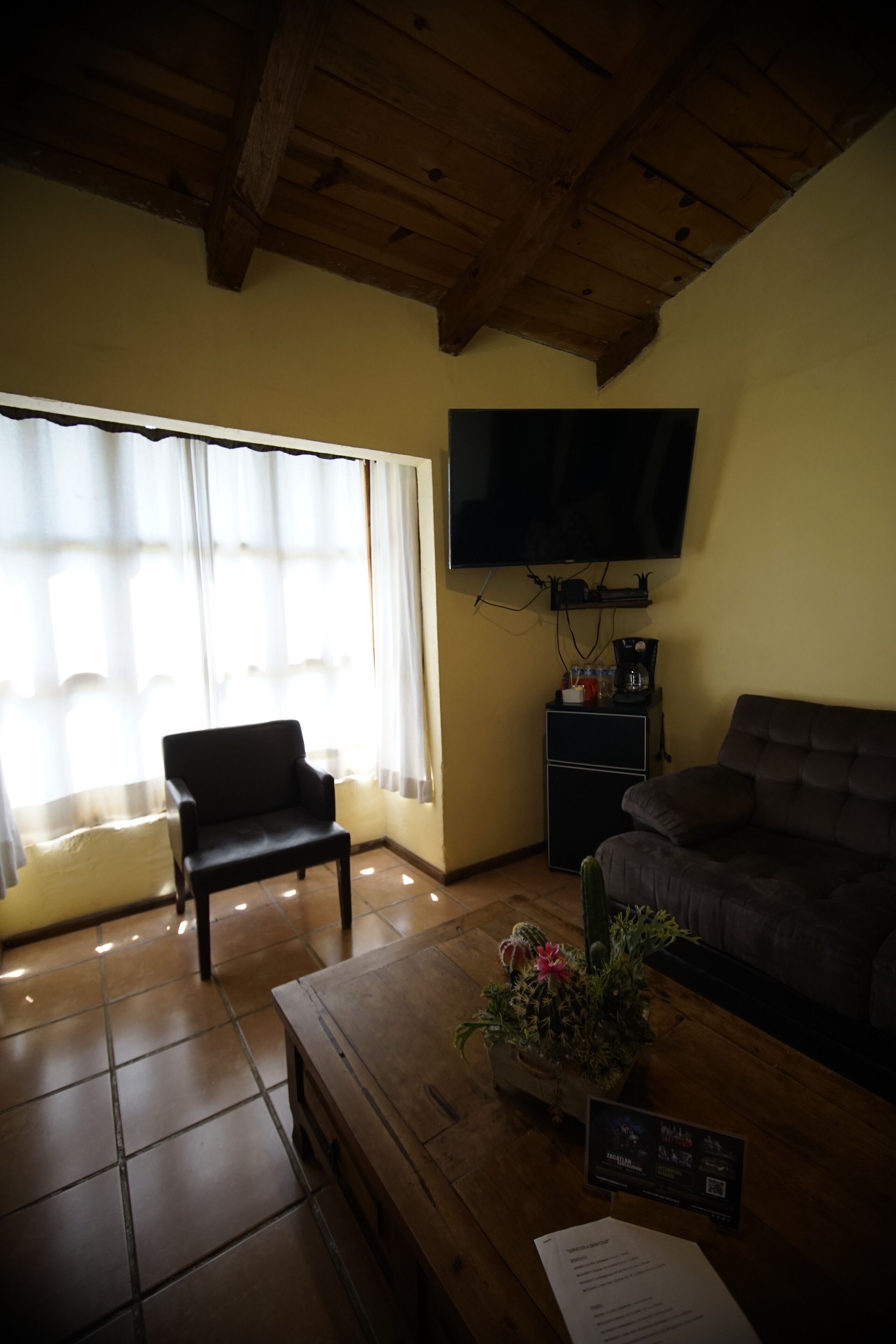 A living room with a wooden ceiling, a TV mounted on the wall, a sofa, a chair, a coffee table with a flower vase, and a small cabinet with a TV stand.