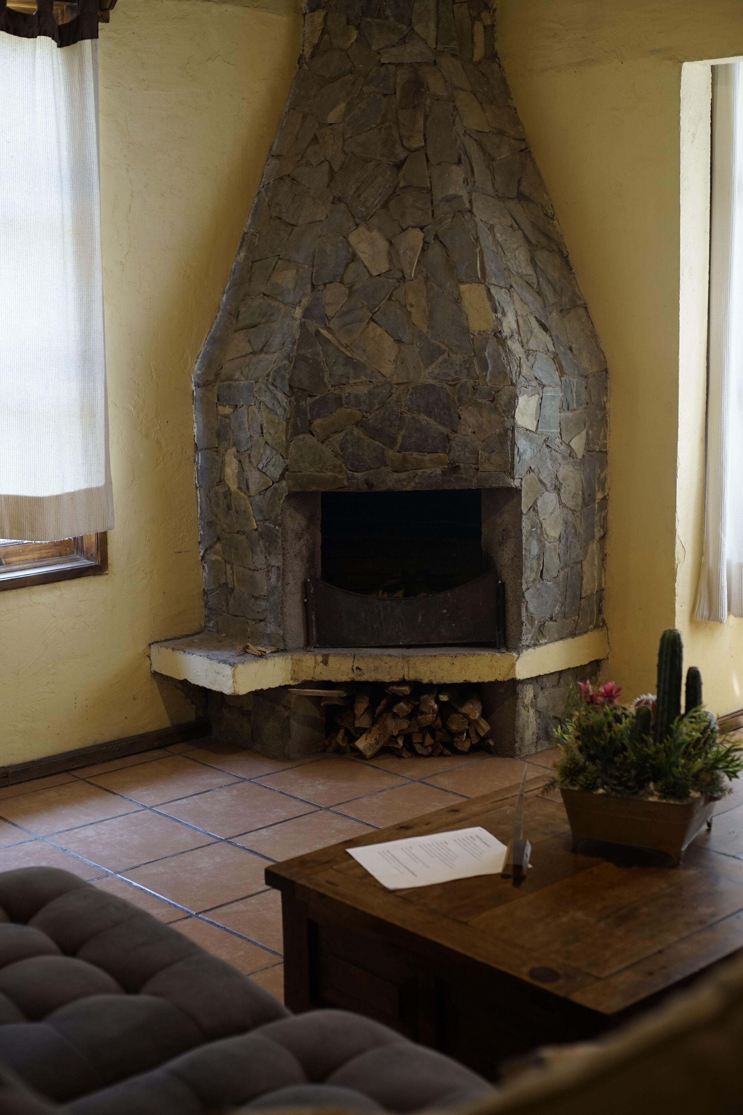 A stone fireplace with a log storage area below, a wooden table with a paper on it, and a potted plant.