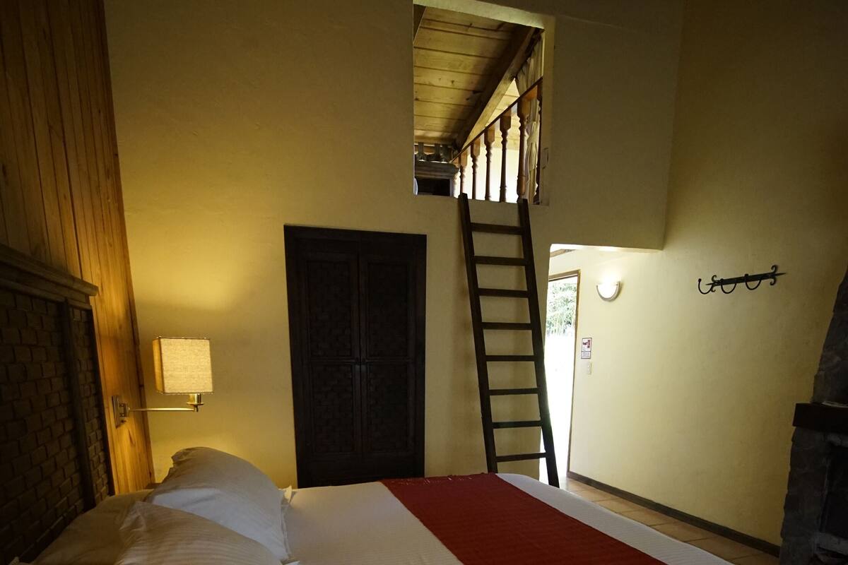 Habitación de hotel con una cama grande, una escalera de madera, una chimenea de piedra y una puerta que da a un balcón.