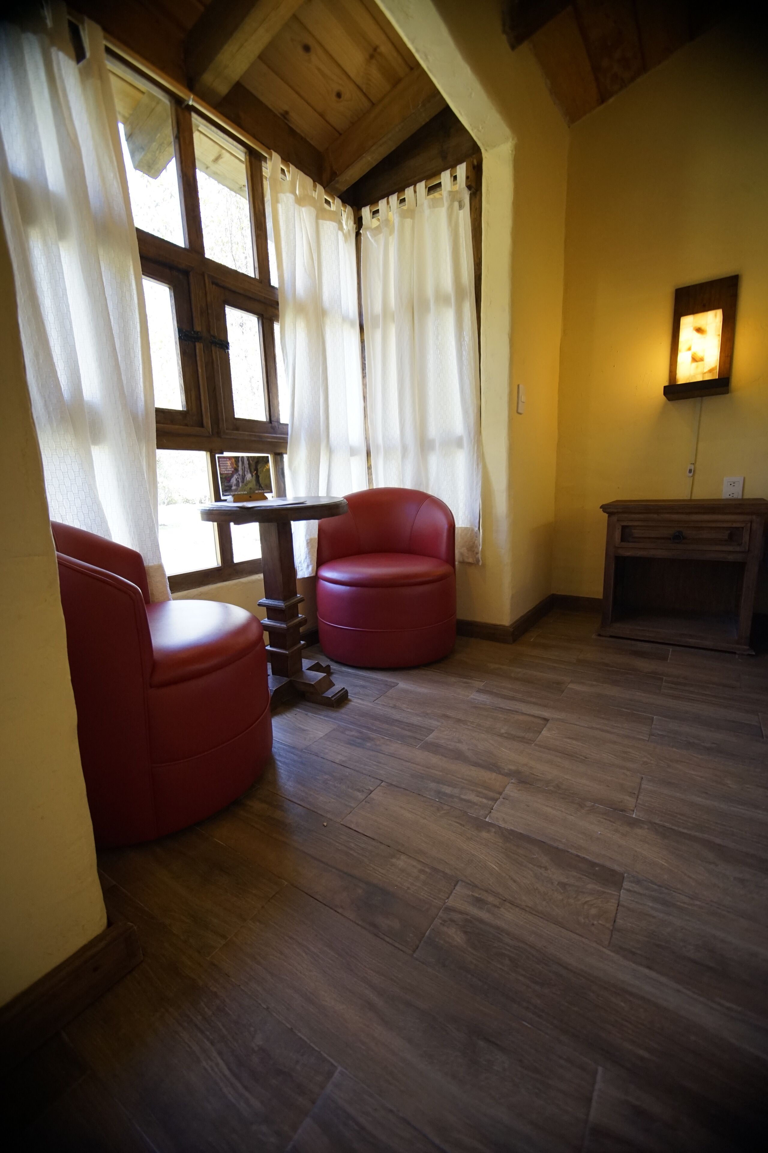 Een kamer met houten vloer, twee rode stoelen, een kleine tafel en een wandlamp.