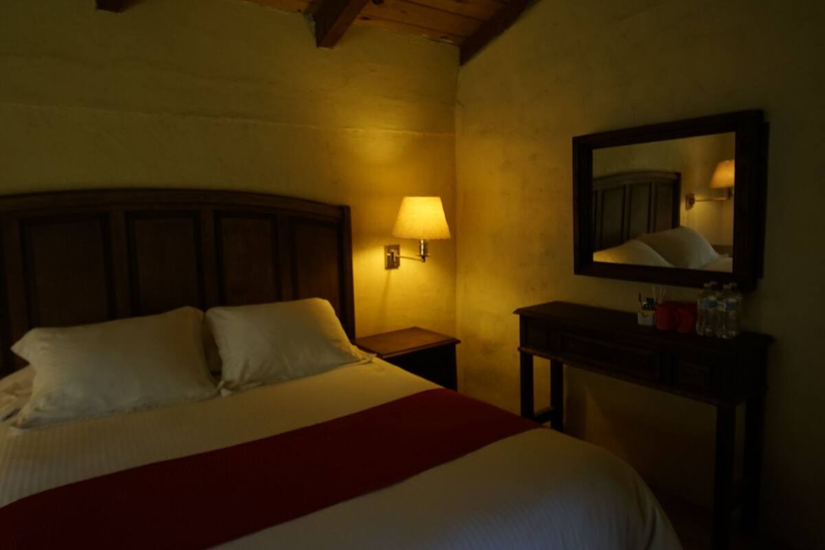 Un hotel habitacion con poca luz, una cama grande, una mesita de noche con una lámpara y un espejo en la pared.