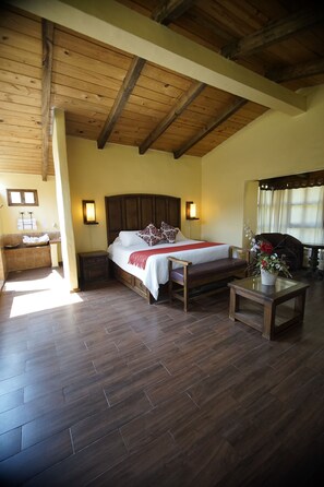 A bedroom with a wooden ceiling, a bed with a white bedspread and pillows, a nightstand, a small table with a vase, and a window with curtains. - Cabañas La Cascada (Zacatlán)