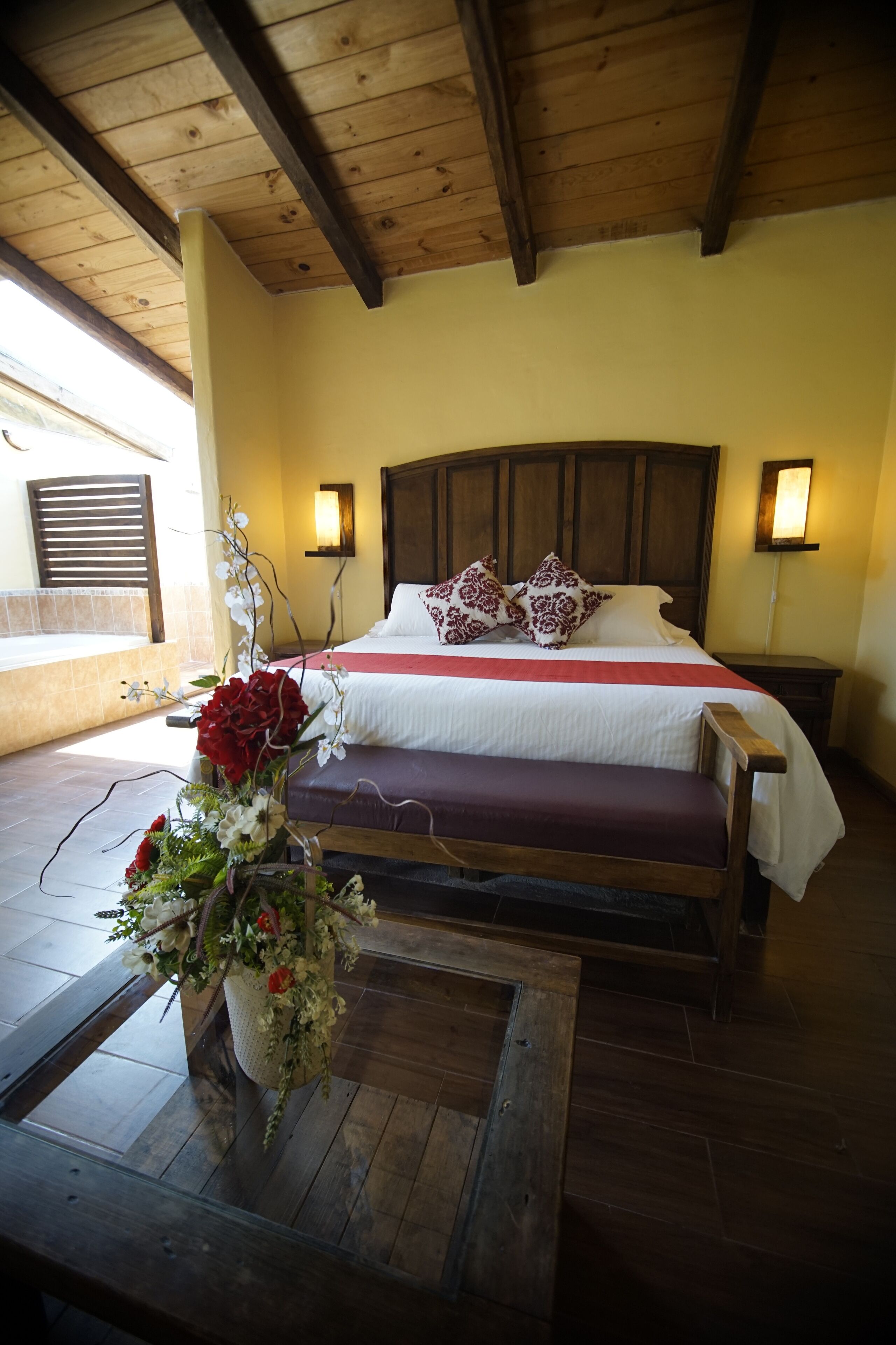 Ein Schlafzimmer mit Holzbalkendecke, einem großen Bett mit weißer und roter Bettwäsche, zwei Nachttischlampen und einer Blumenkomposition auf einem Tisch.