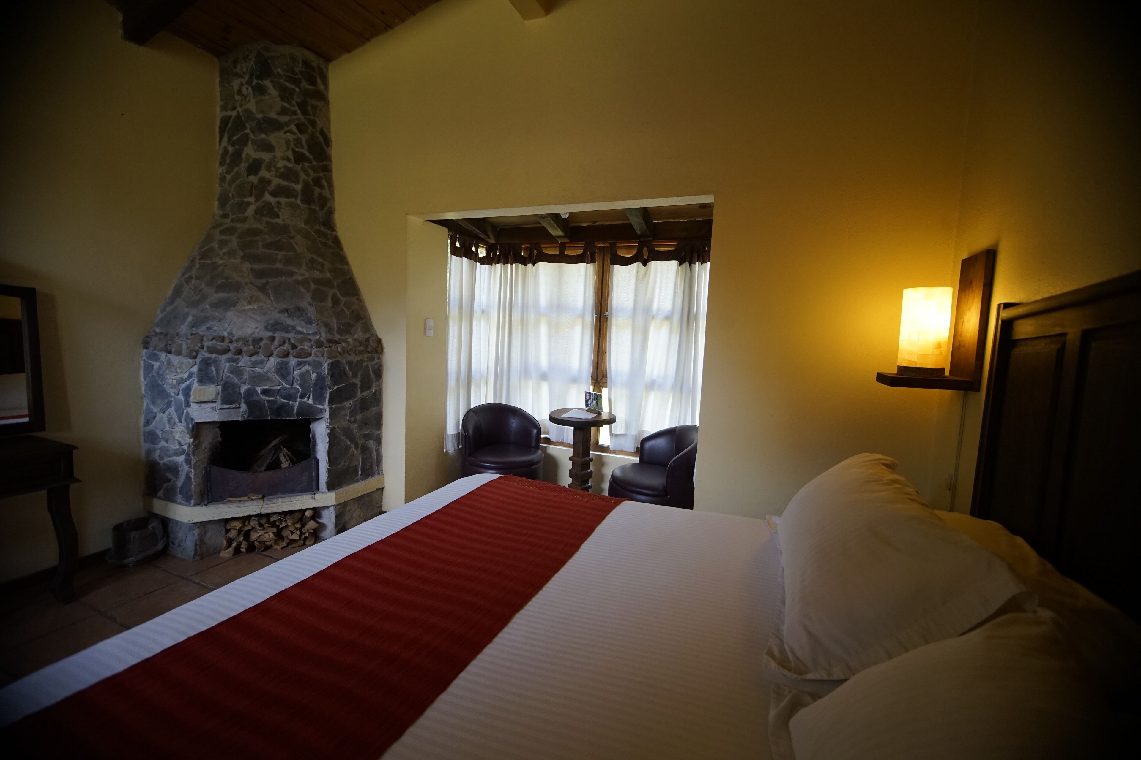 Una habitación con chimenea de piedra, una cama con colcha roja y blanca, dos sillas, una mesita y una ventana con cortinas.