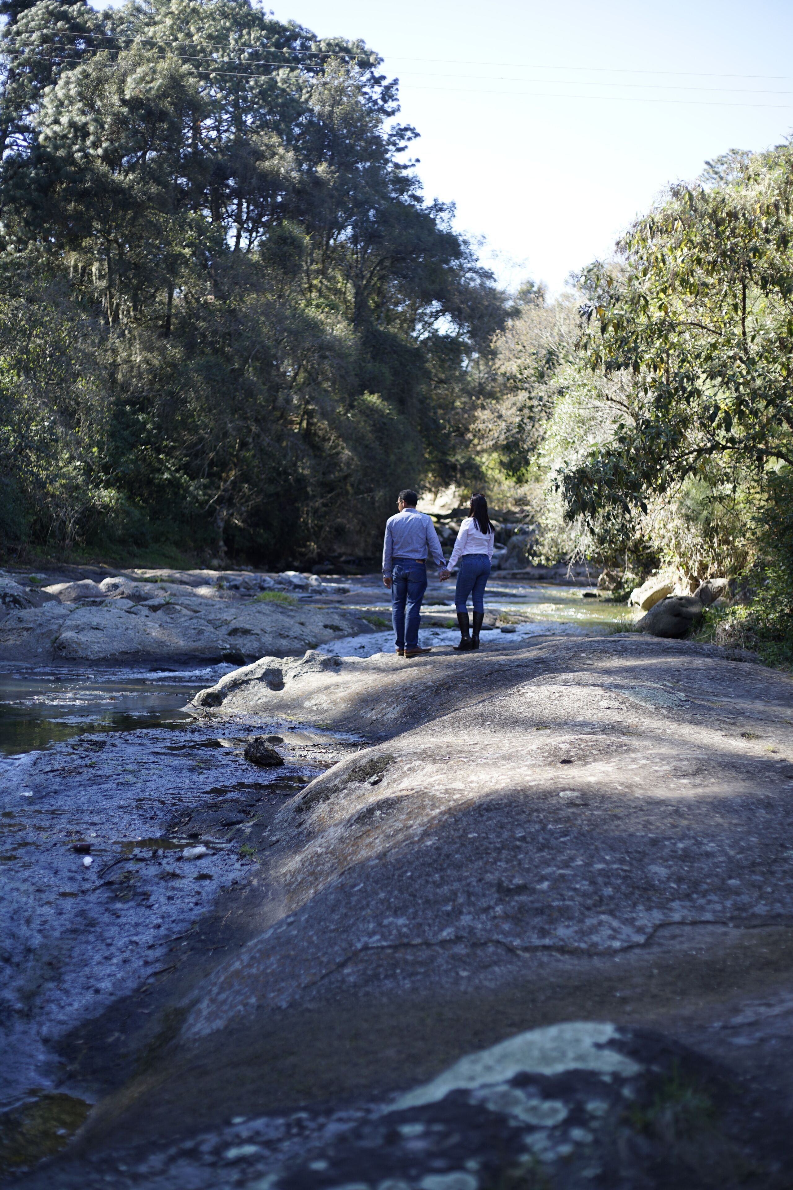 A couple walking on a rocky path beside a river in a wooded area.