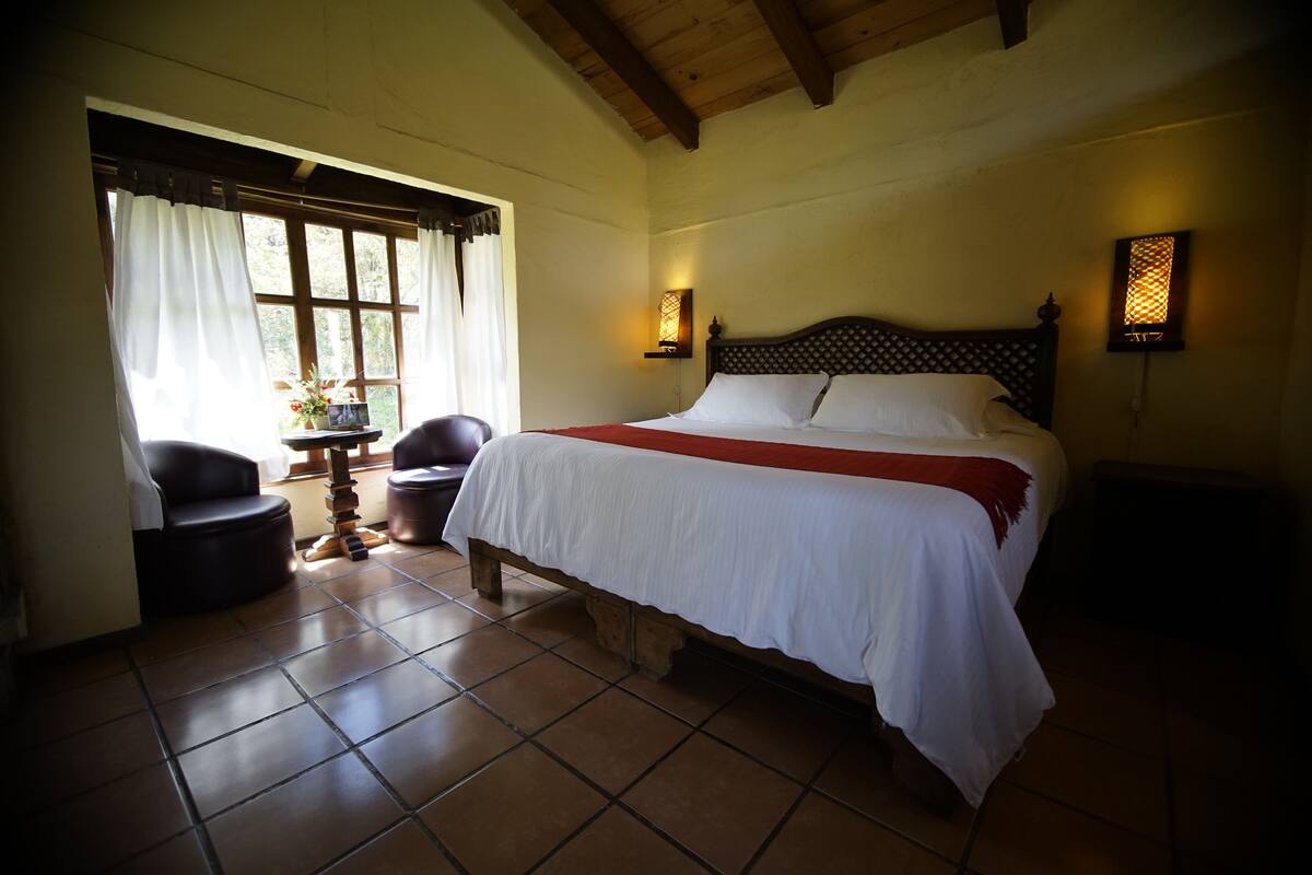 Un dormitorio con una cama grande, una silla, una mesita y una ventana con cortinas.