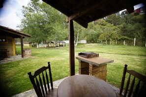 A patio area with a round table and two wooden chairs, a brick barbecue grill, and a covered seating area in a backyard with a grassy lawn and trees. - Cabañas La Cascada (Zacatlán)
