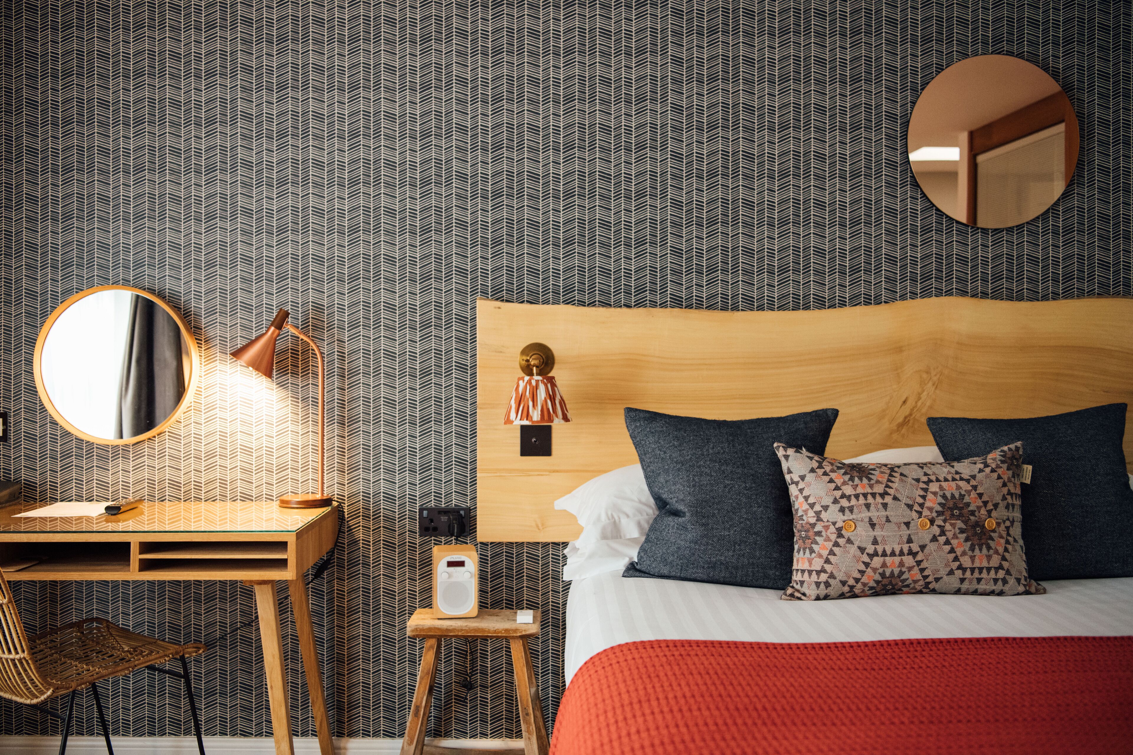 A neatly arranged bedroom with a wooden desk, a chair, a bedside table with a lamp, and a bed with patterned pillows.
