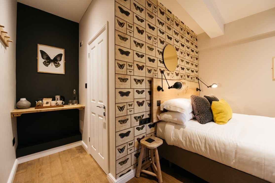 Un dormitorio con cama, mesita de noche, taburete, espejo adosado a la pared y una pared decorada con reproducciones de mariposas.