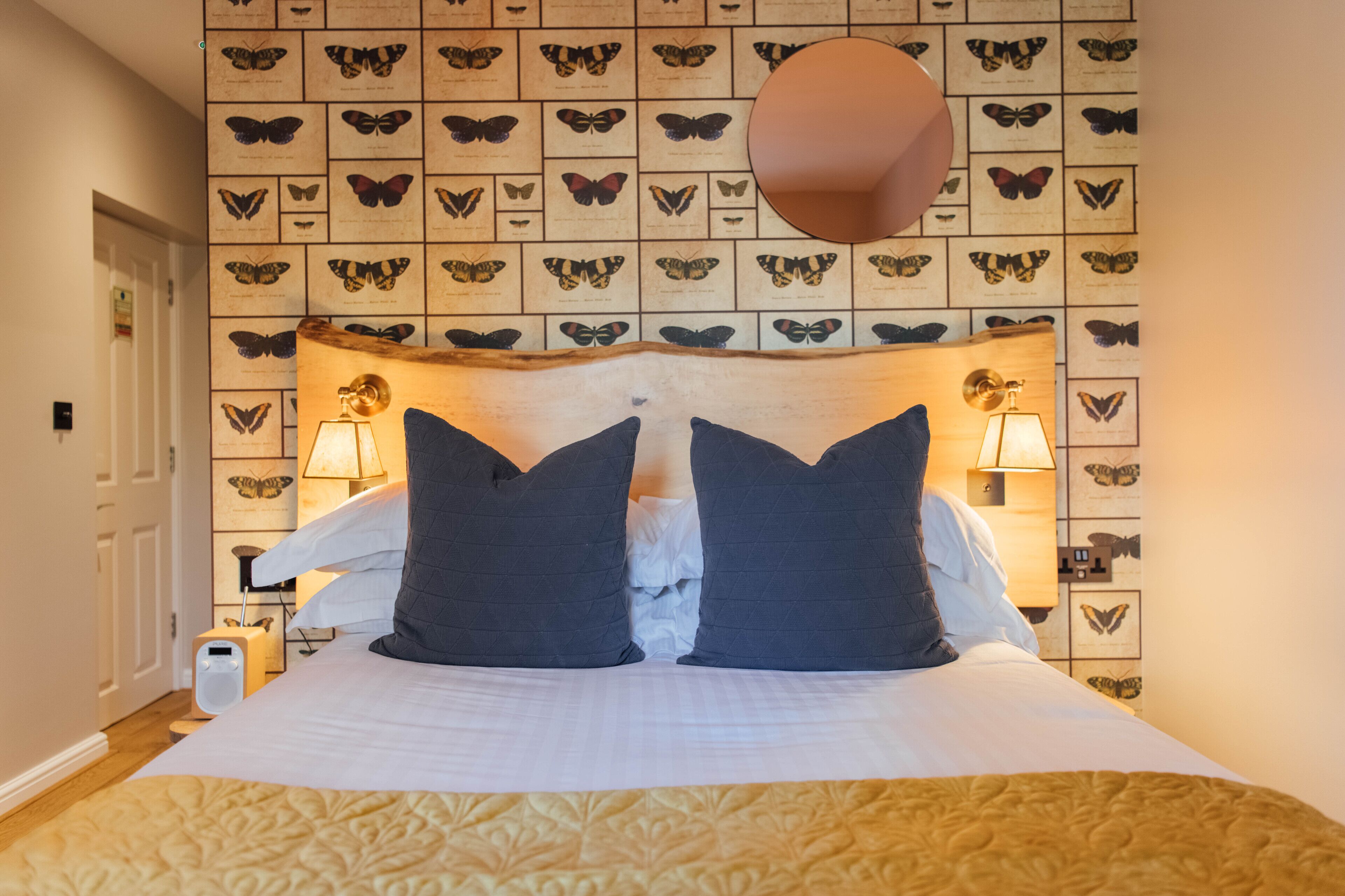 Una habitación de hotel con una cama, dos lámparas de noche, una pared llena de estampas de mariposas y un cabecero de madera.