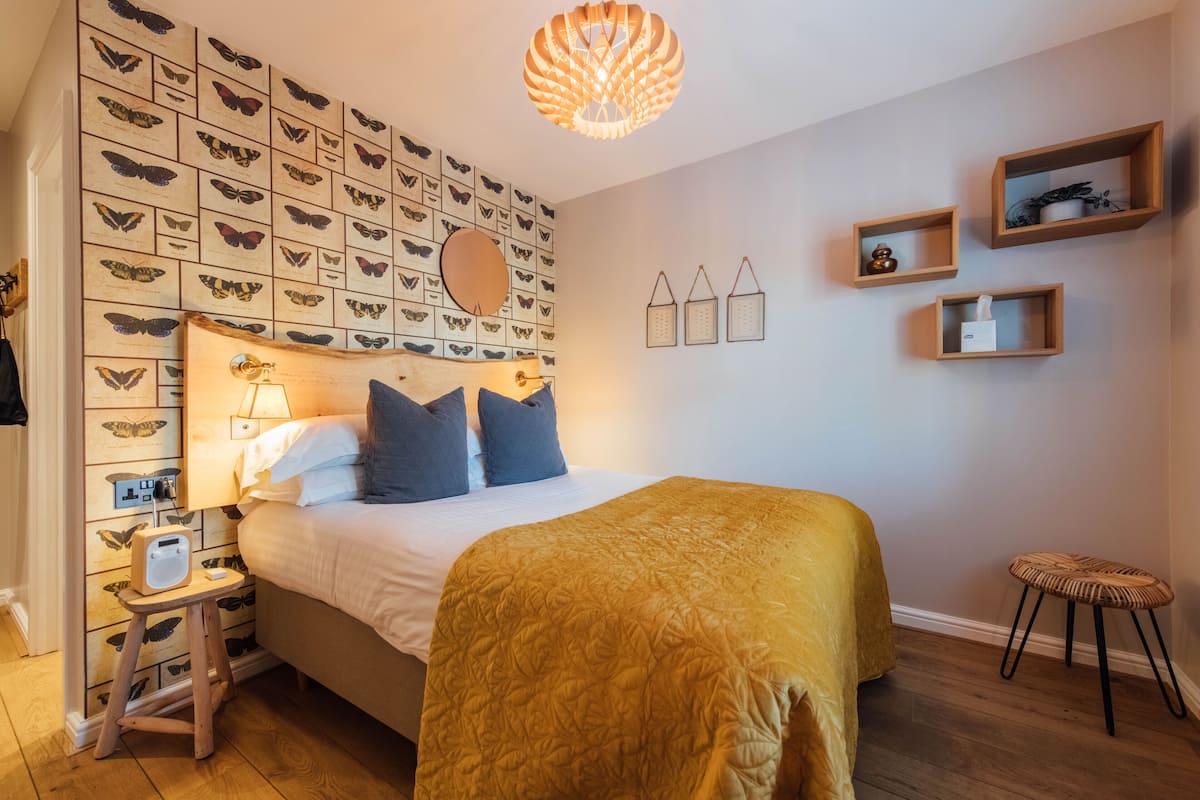 A bedroom with a bed, a wall-mounted butterfly display, a bedside lamp, and a small stool.