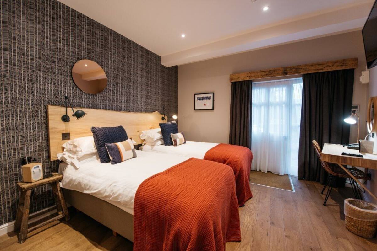 A hotel room with two beds, a wooden headboard, a desk with a chair, a mirror, and a window with curtains.