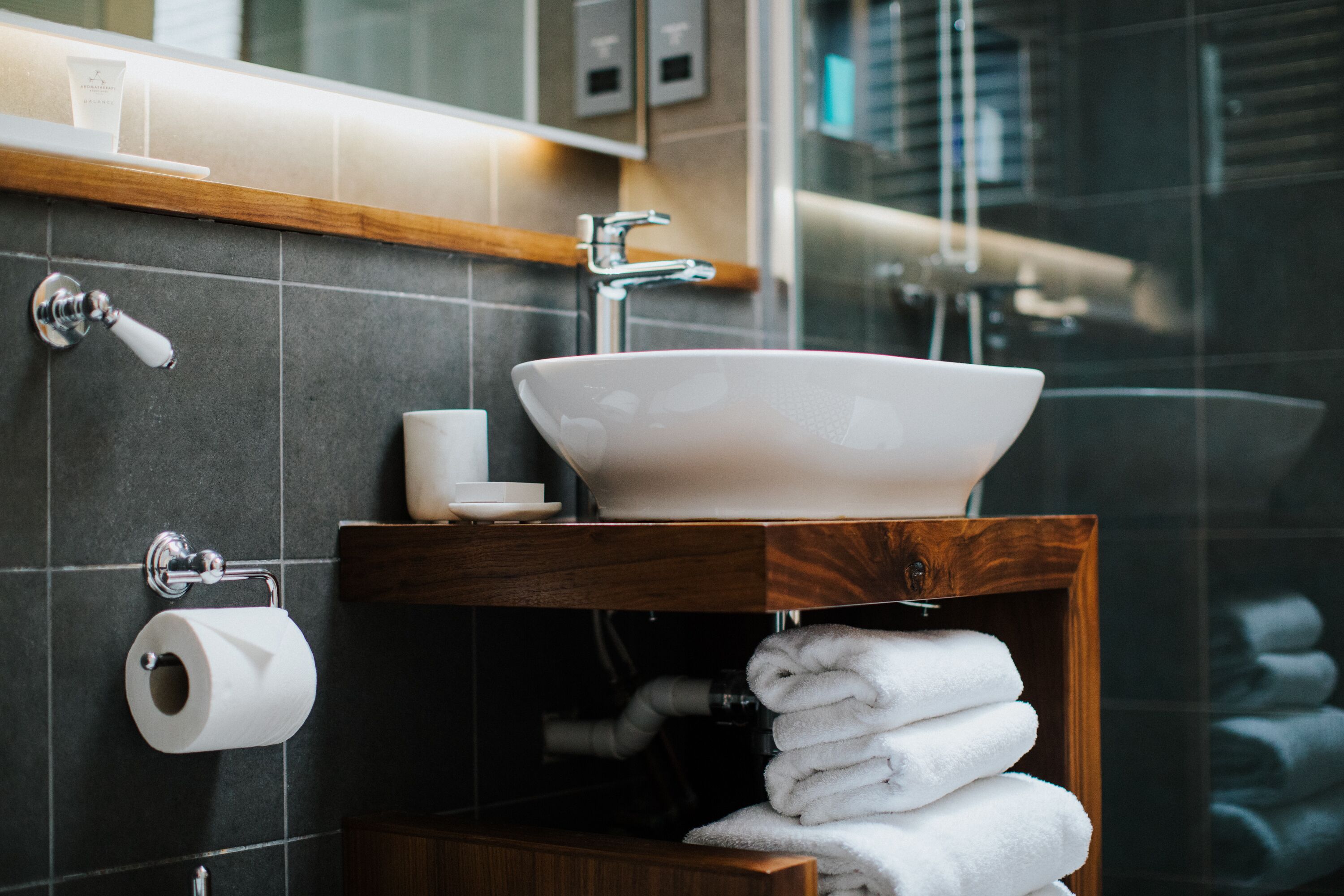 Un baño con un lavabo blanco, un tocador de madera, un espejo, un perchero para toallas y un dispensador de papel higiénico.