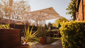 An outdoor patio area with a large umbrella, potted plants, and a wooden bench. - The Fox at Willian  (Letchworth)