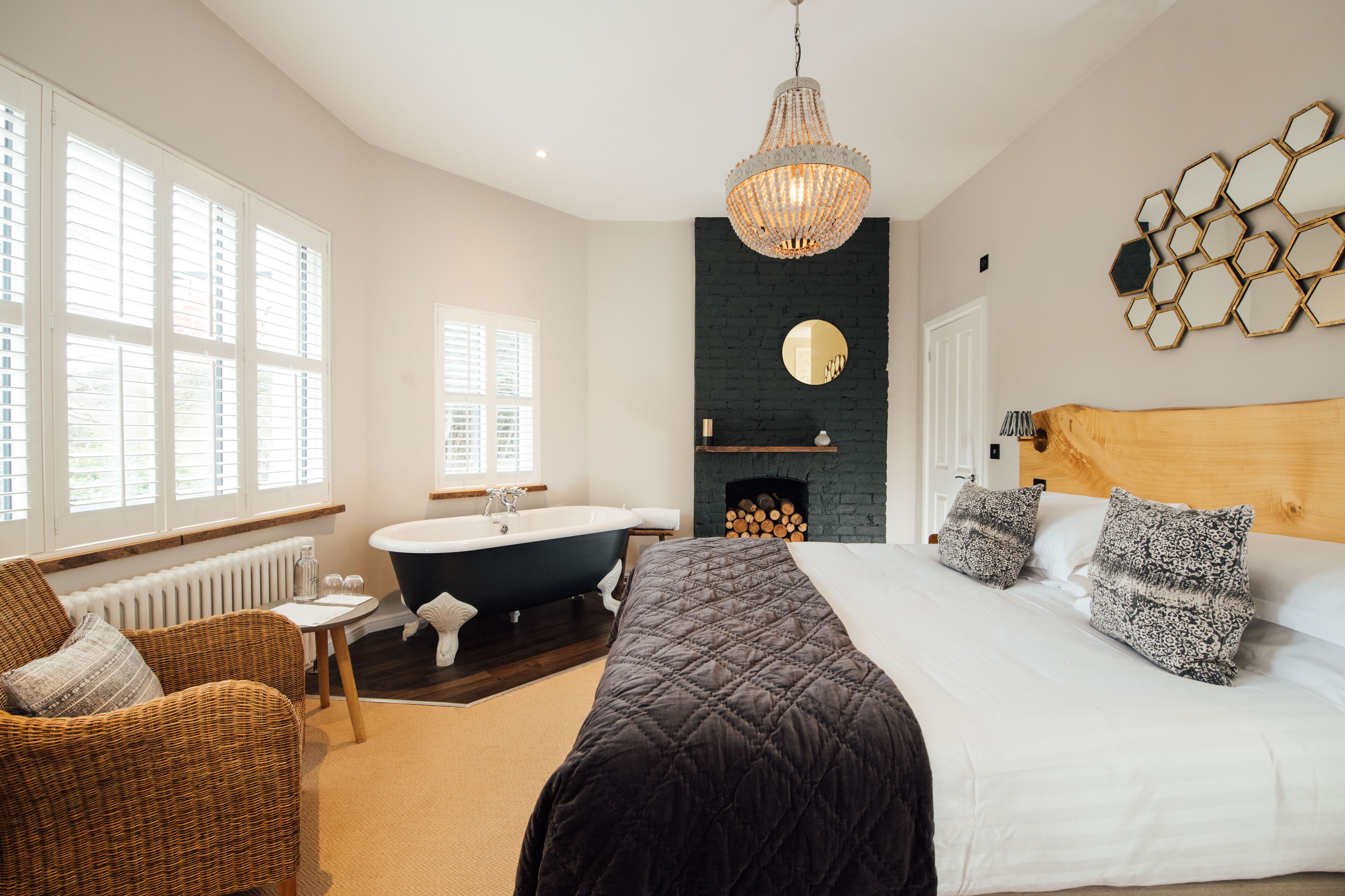 A modern bedroom with a large bed, a wooden headboard, a black fireplace, a freestanding bathtub, and a wicker chair.