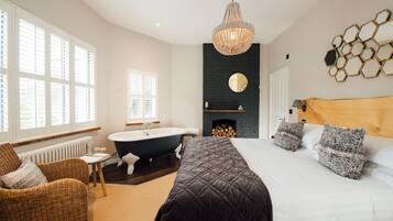 A modern bedroom with a large bed, a wooden headboard, a black fireplace, a freestanding bathtub, and a wicker chair.