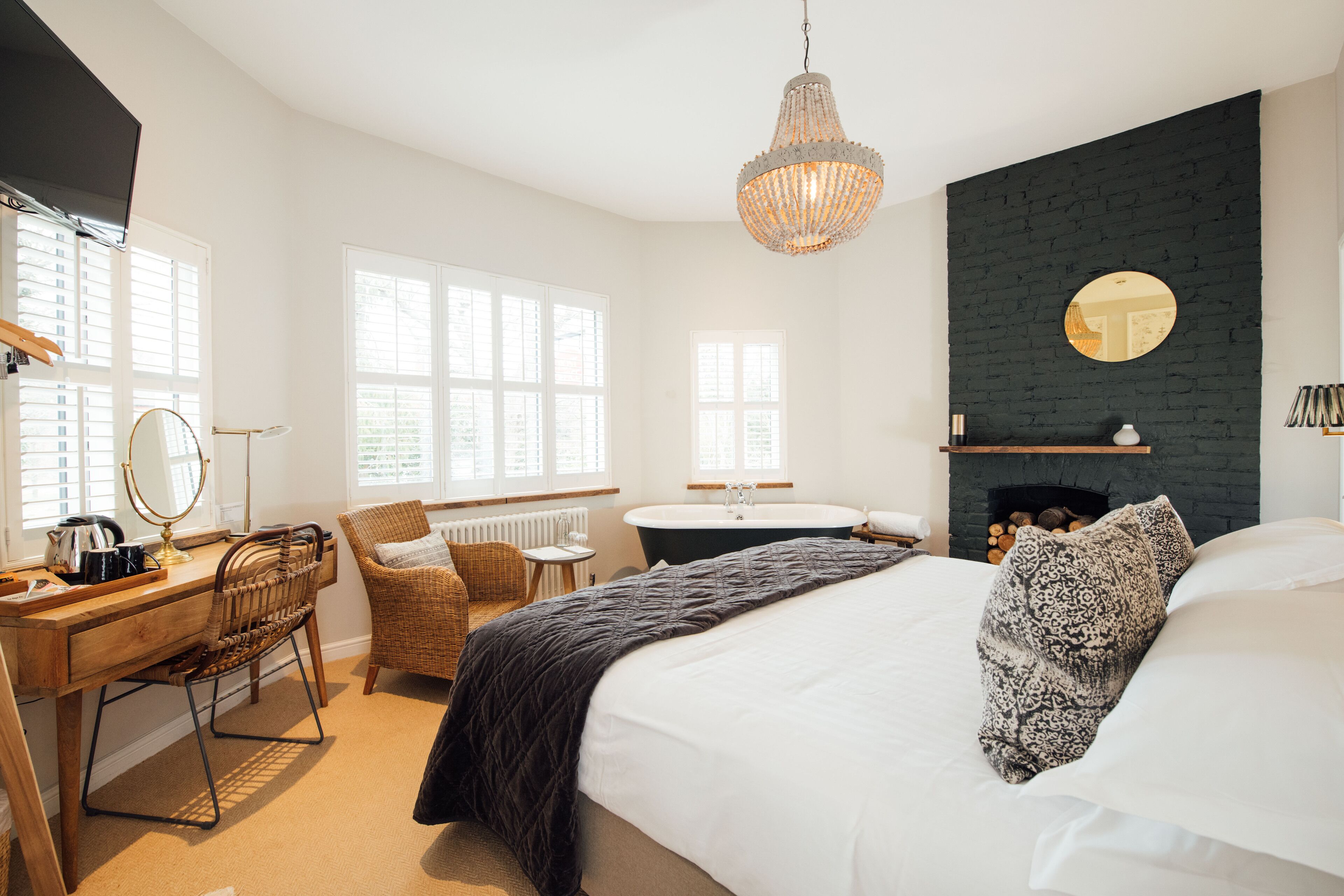 A bedroom with a bed, a desk, a chair, a round mirror, a chandelier, a fireplace, and large windows.