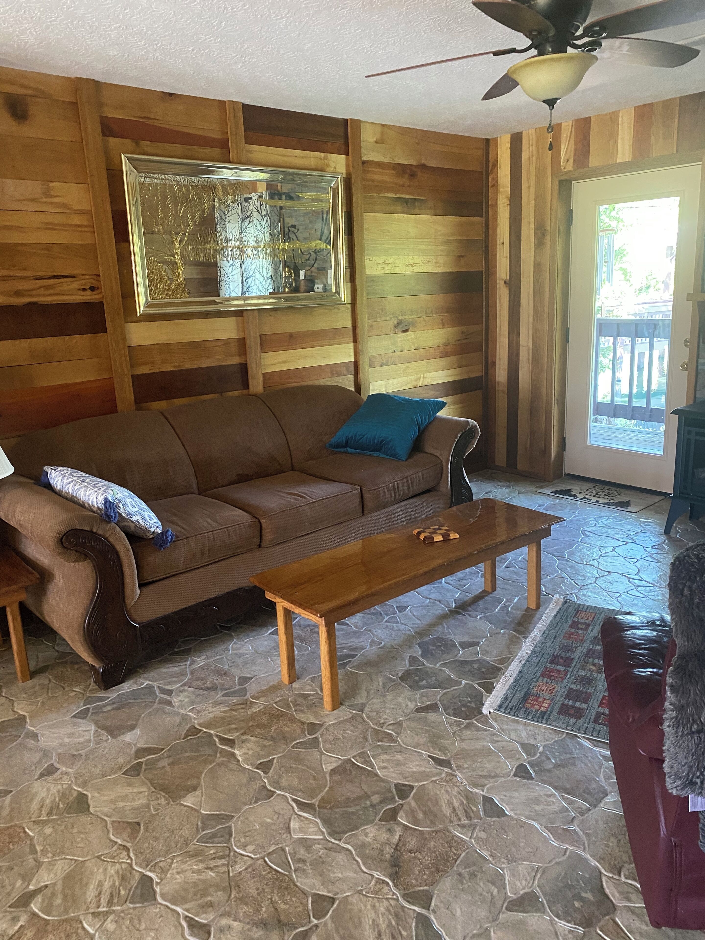 A wooden interior with a stone floor, a brown sofa, a wooden coffee table, and a ceiling fan.
