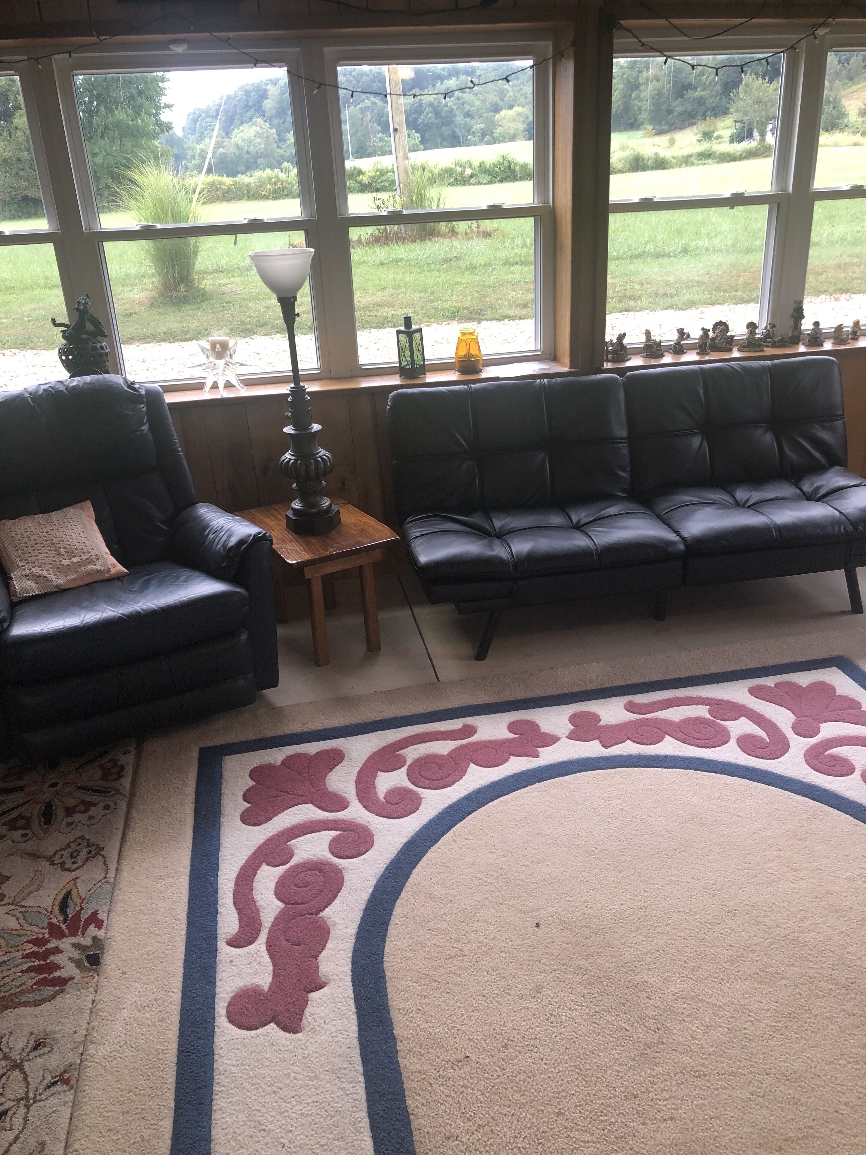 Ein Zimmer mit einer schwarzen Ledersofa, einem schwarzen Ledersessel, einem kleinen Tisch mit einer Lampe und einem großen Fenster mit Blick auf Grünflächen.