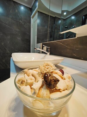 A modern bathroom with a large mirror, a white sink, and a glass bowl containing various bath items on the countertop.
