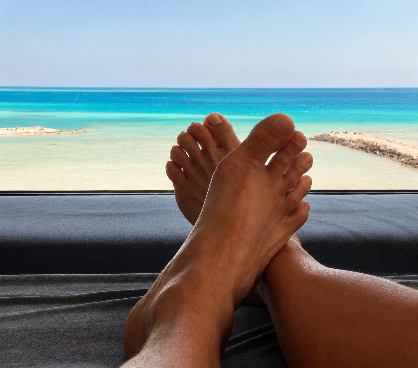 Voeten rusten op een donkere ondergrond, met uitzicht op een strand en de oceaan door een raam.