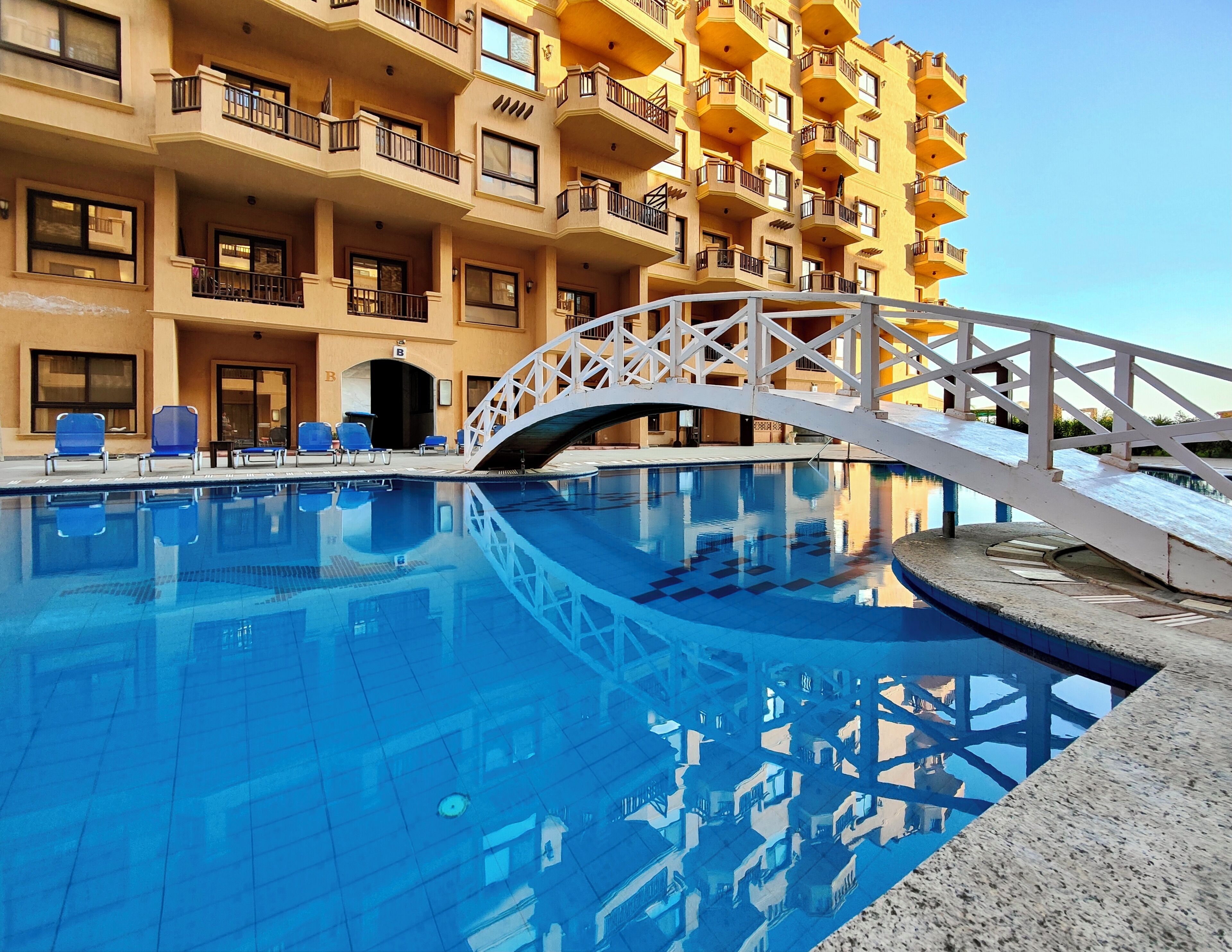 A swimming pool with a white bridge, surrounded by a multi-story building with balconies and a sign indicating room B.