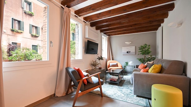Un salon moderne avec un canapé, un fauteuil et un pouf, un plafond en bois et une vue sur un bâtiment à l’extérieur.