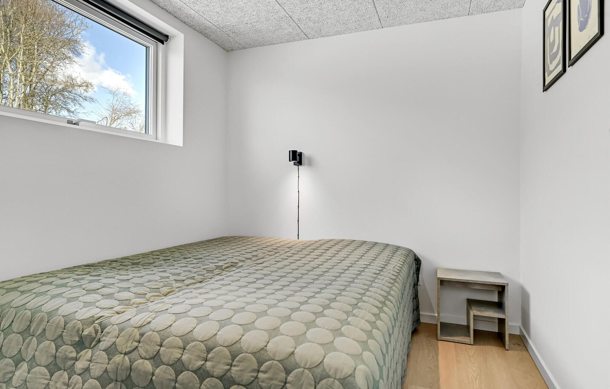 A bedroom with a bed, a bedside table, a wall-mounted light, and a window.