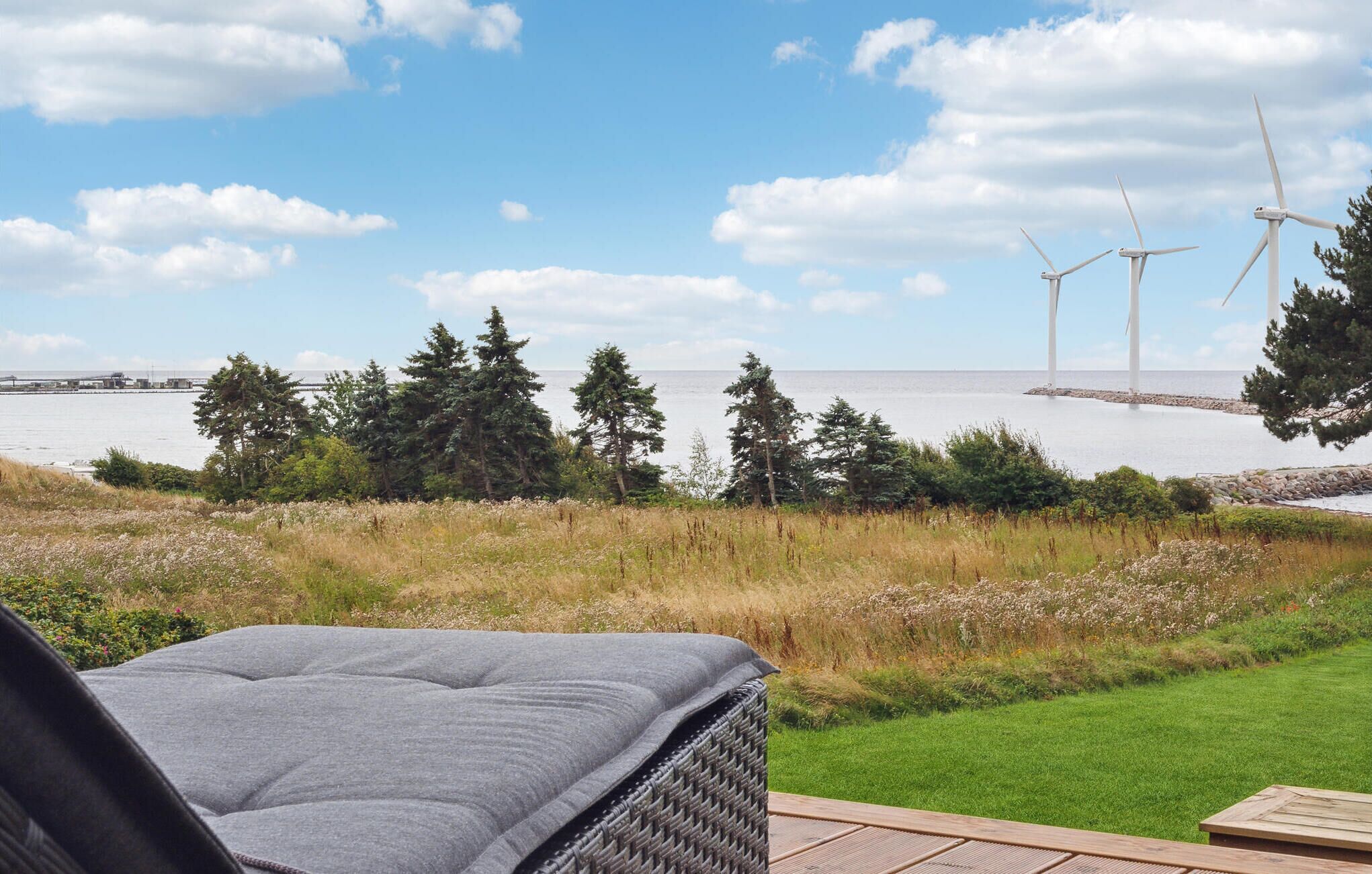 Un patio con una silla de mimbre, una mesa de madera y vista al mar, con aerogeneradores a lo lejos.