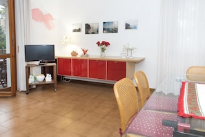 Un coin repas avec une table en verre, des chaises en osier, un buffet rouge et un téléviseur posé sur un meuble.