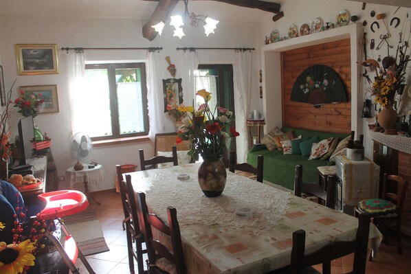 Un coin repas avec une table dressée pour quatre personnes, entourée de chaises en bois. Un vase rempli de fleurs est posé sur la table, et un ventilateur se trouve sur une petite table à côté de l’espace repas.