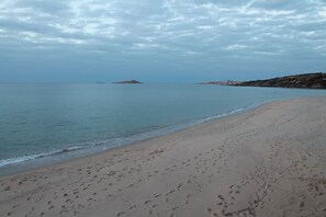 En strand med fodaftryk i sandet, roligt vand og fjerne landformer under en overskyet himmel.