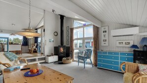 A spacious living room with a wooden table, a blue dresser, wicker furniture, a fireplace, and large windows.