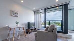 A modern living room with a sofa, a dining table with chairs, a TV, and a large window offering a view. - City Escape 1-Bedroom Studio with Pool in CBD (Auckland)