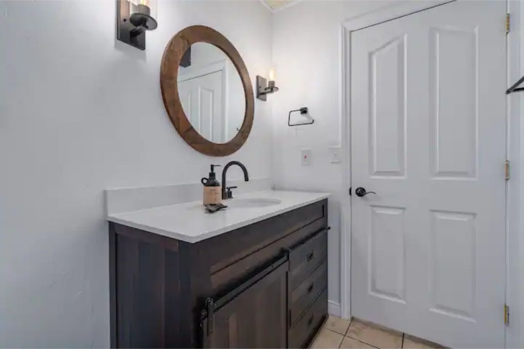 Un baño con lavamanos blanco, tocador oscuro y un espejo redondo.
