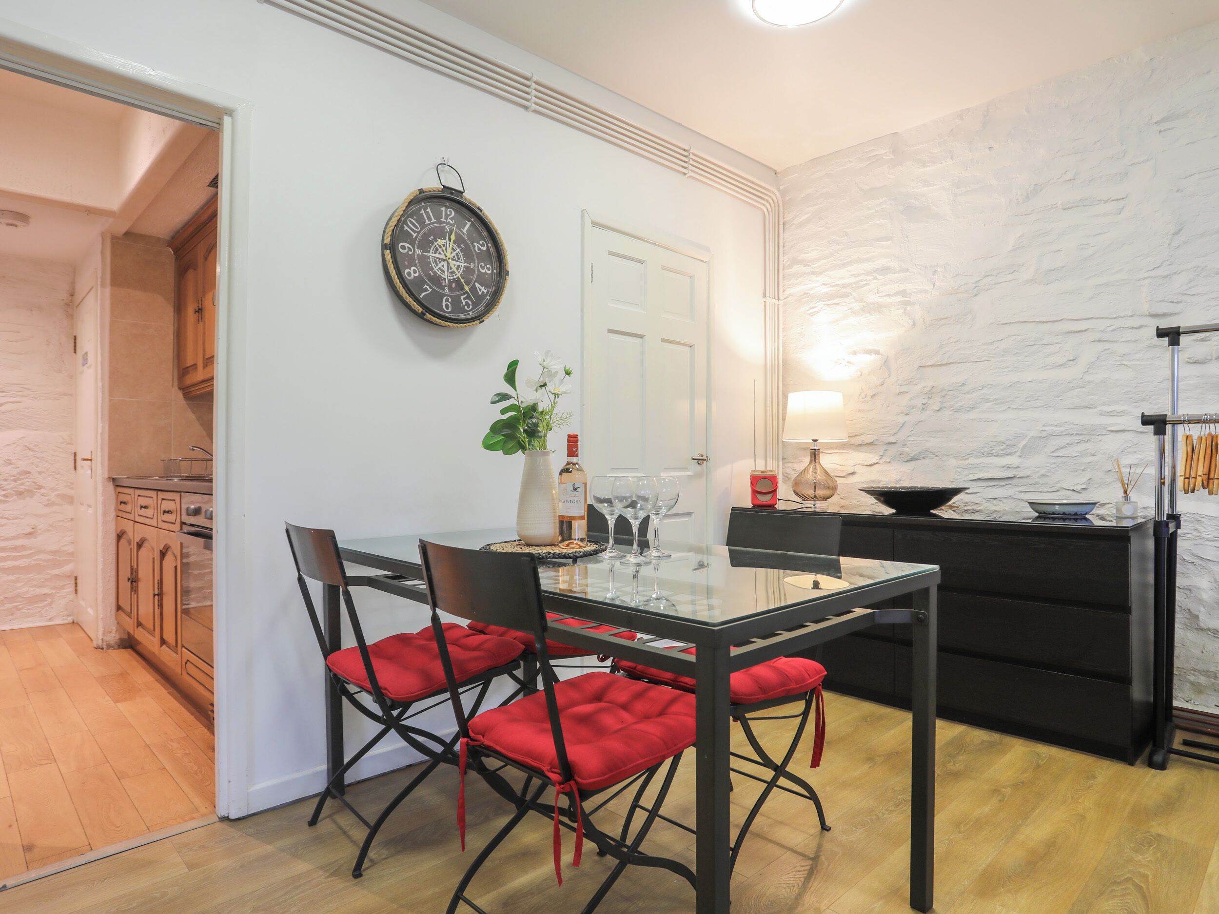 A dining area with a glass table, red cushioned chairs, a wall clock, a vase with flowers, and a wine bottle.