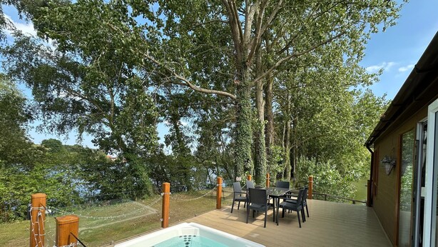 A hot tub on a wooden deck with a table and chairs, surrounded by trees and a view of a body of water.