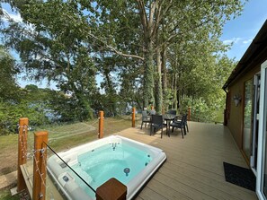 Un jacuzzi en una terraza de madera con una mesa y sillas, rodeado de árboles y vistas a un cuerpo de agua.