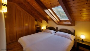 Une chambre avec un plafond en bois, une lucarne, un lit avec du linge de lit blanc, une table de chevet avec une lampe et une armoire.