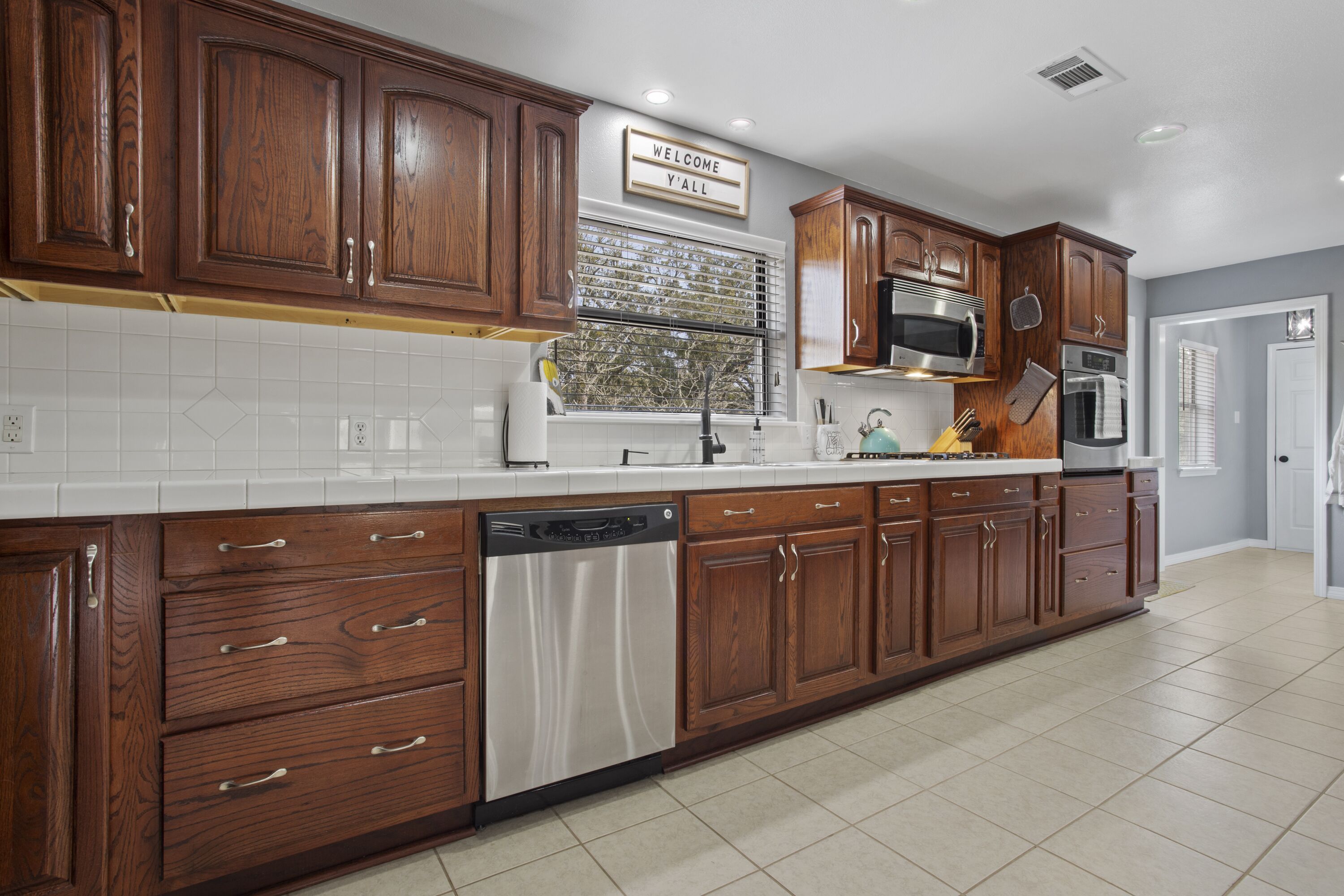 A kitchen with wooden cabinets, a dishwasher, a microwave, and a sign that says "WELCOME Y'ALL".